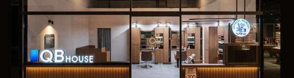QB House express haircut storefront seen through glass, showing interior booths and bright signage at night outside.