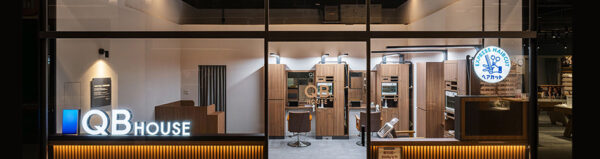 Night view of the QB House hair salon storefront with glass walls, warm lighting, and wooden styling stations inside.