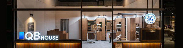 Storefront of QB House hair salon at night; glass windows show a reception area, wooden styling stations, and an illuminated QB House sign with an Express Hair logo