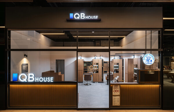 Storefront of QB House hair salon with glowing logo and wood-paneled interior visible through glass doors at night
