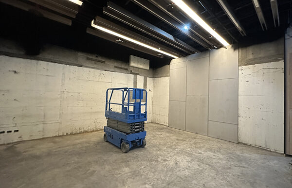 Blue scissor lift parked in a bare concrete-room with exposed ceiling lights and walls nearby.