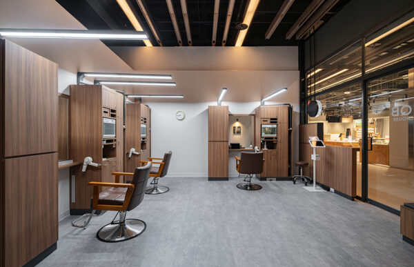 Modern salon interior with row of wooden styling stations, brown chairs, and built-in appliances under bright ceiling lights.