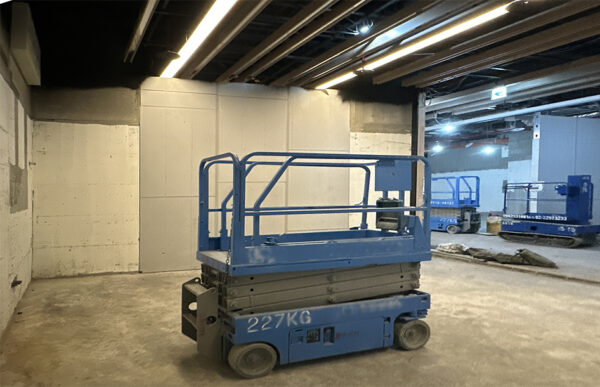 Blue scissor lift parked in an unfinished indoor construction area with concrete walls and exposed ceiling beams.