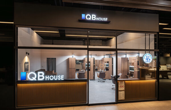 Storefront of QB House hair salon with glass facade and illuminated logo, visible interior with styling stations and warm wooden accents.