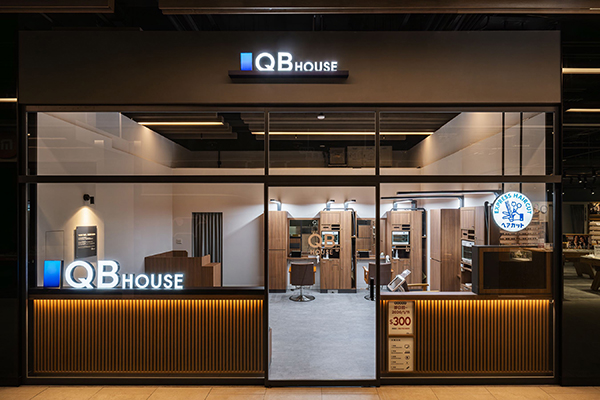 Exterior of a QB House storefront with glass walls, illuminated logo, and warm interior lighting inside a mall.